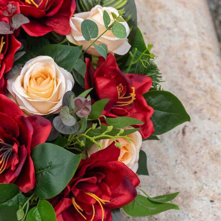 Stroik na cmentarz ze sztucznych kwiatów. Czerwony amarylis, różowe róże. Detal kompozycji. Kwiaty na tle jasnej płyty nagrobnej.